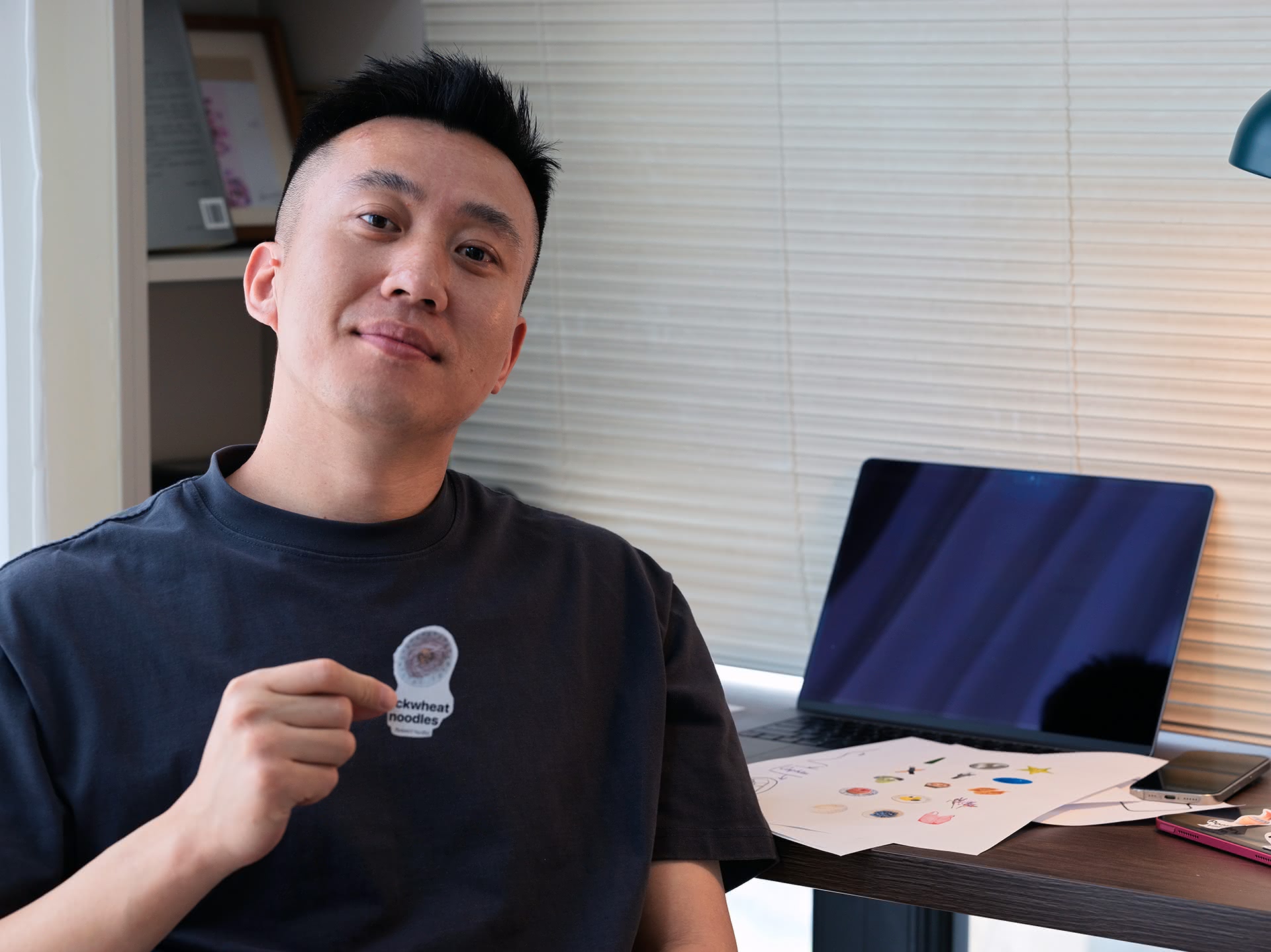 A man sits indoors at a desk holding up a sticker that shows an image of buckwheat noodles and contains the words. On the desk is a MacBook and a series of papers with stickers on them.